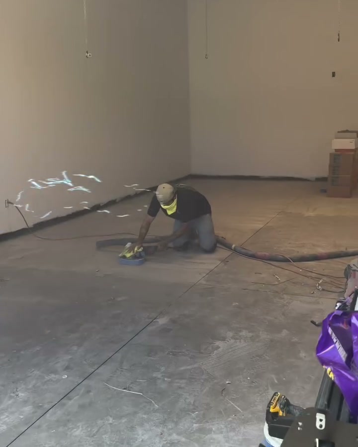 A Ghost Ridge Epoxy Co. installer kneeling on a bare shop slab, grinding the concrete with a vacuum hose running behind him.