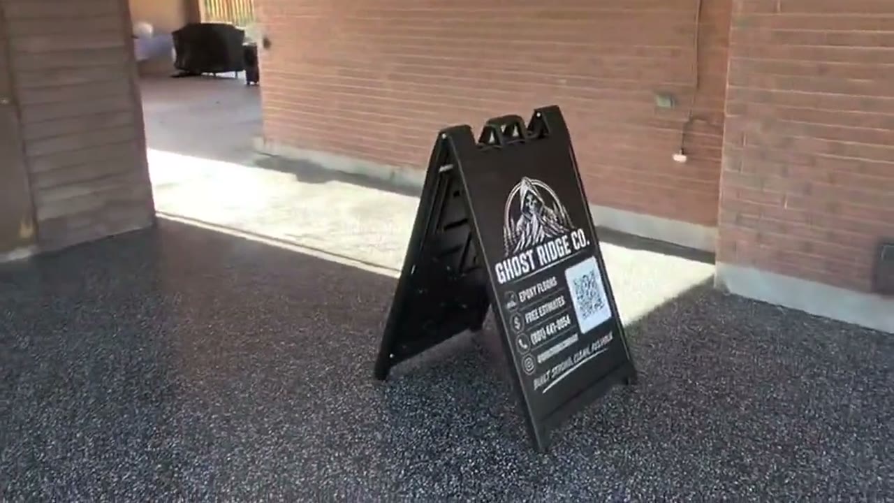 The Ghost Ridge Epoxy Co. yard sign standing on a finished raven flake carport floor beside a brick wall.