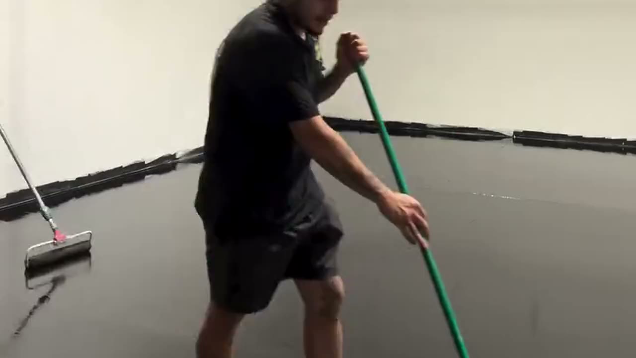 An installer rolling a wet epoxy base coat across a large commercial shop floor, the wall reflected in it.