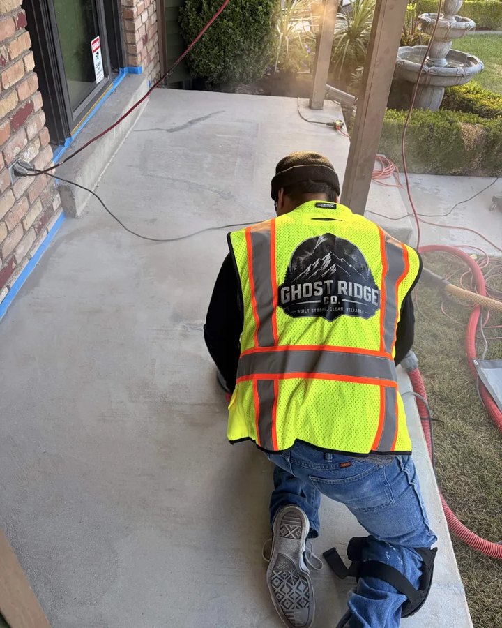A Ghost Ridge Company LLC installer in a branded hi-vis vest, kneeling on a front porch slab part way through grinding it.